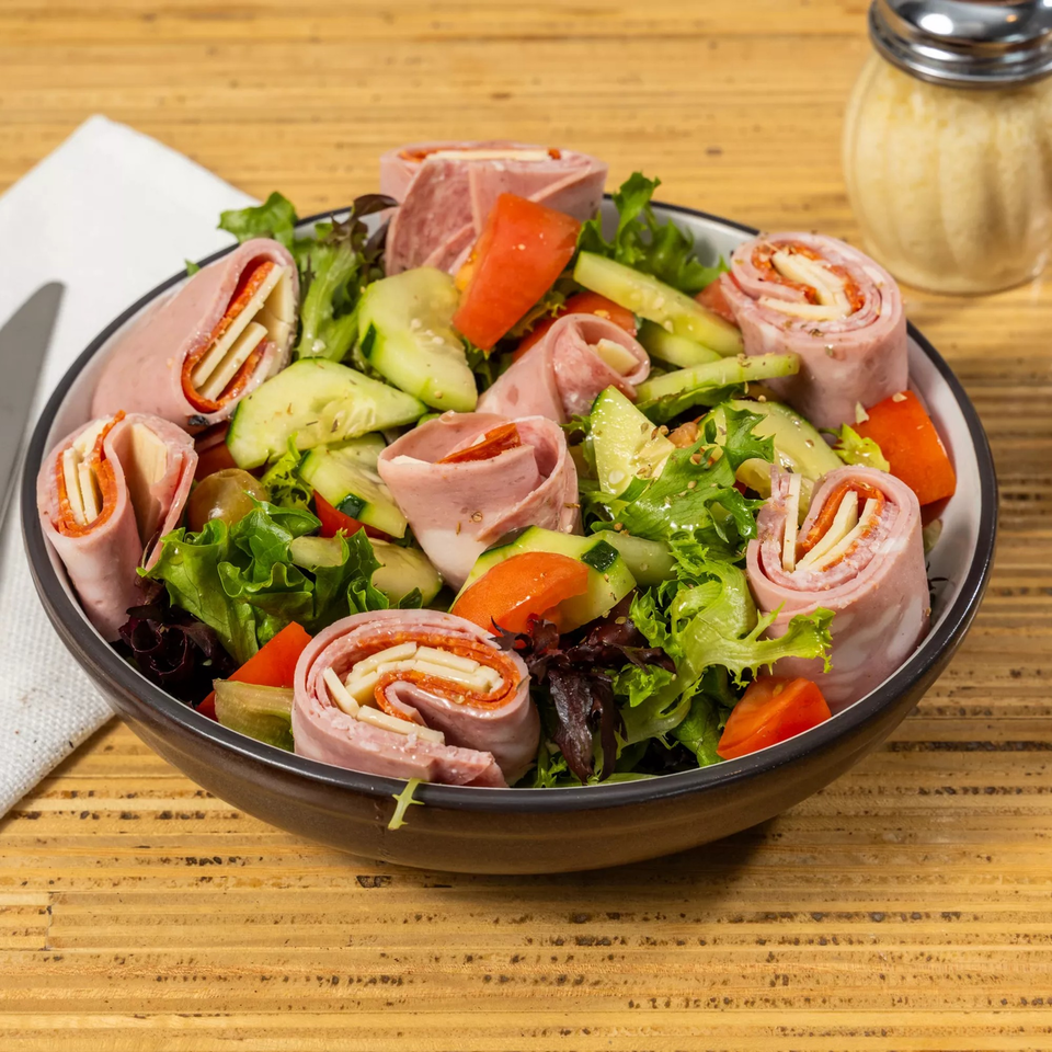 A fresh salad with mixed greens, cucumbers, tomatoes, and rolled deli meat with cheese and pepperoni.