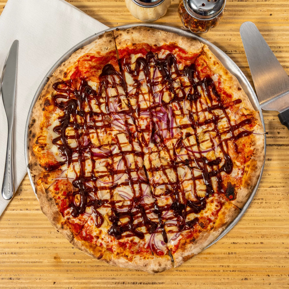 BBQ chicken pizza with red onions and a drizzle of sauce, on a wooden table with utensils.