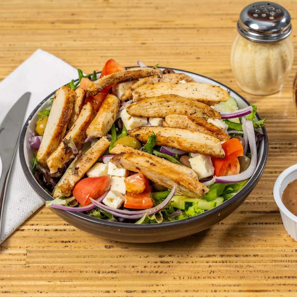 Grilled chicken salad with tomatoes, feta, olives, and red onion on a wooden table.